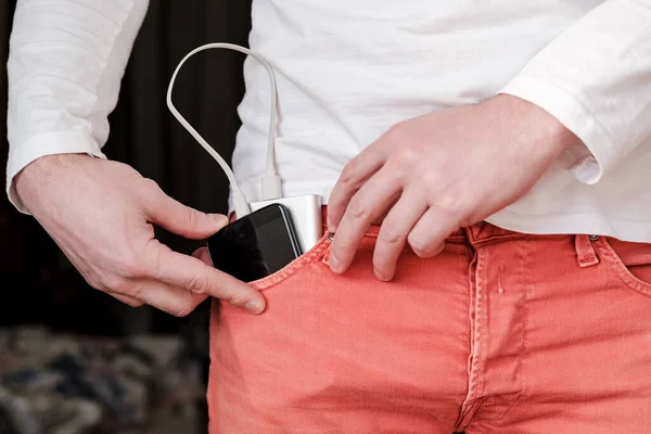 Männliche Hand steckt die Powerbank und das daraus aufladende Handy in die Vordertasche korallenfarbener Jeans. — Stockfoto