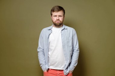 Hüzünlü, üzgün, sakallı bir adam yeşil arka planda kot pantolon ve somurtkan ceplerine ellerini koydu..