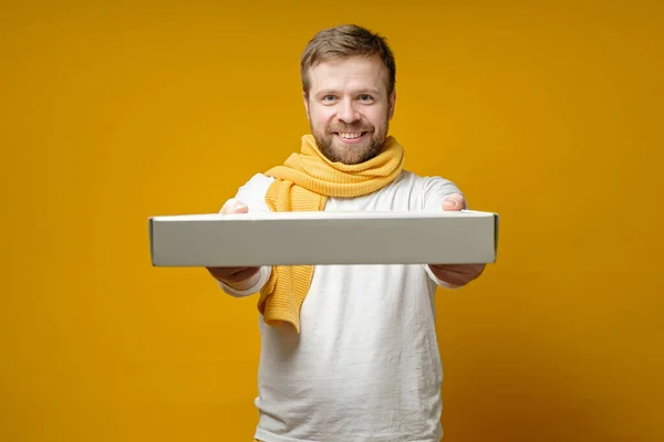 Pleased man wearing a scarf came to a meeting with friends with a pizza, he happily smiling and holding out a box.