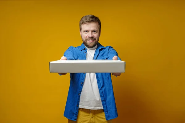 El hombre feliz con una camisa azul llegó a una reunión con amigos con una pizza, sonríe y sostiene una caja, sobre un fondo amarillo . — Foto de Stock