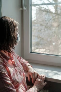 Tıbbi maskeli üzgün kadın pencerenin kenarında oturur ve dışarı bakar. Virüs salgını ve salgın sırasında karantinadadır..