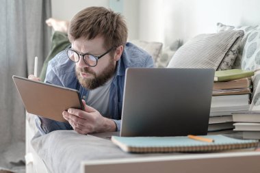 Öğrenci uzaktan çalışıyor, elinde tablet, dizüstü bilgisayar ve kitaplarla kanepede yatıyor. Karantina sırasında çevrimiçi eğitim kavramı.