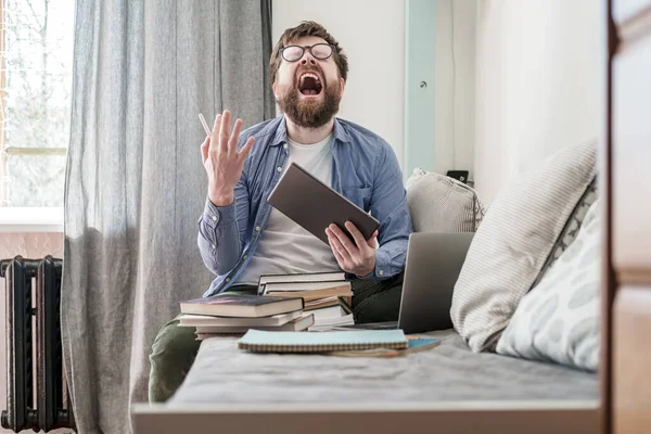 Studerande i stress, han håller en tablett och skriker, sitter på en soffa, hemma. Begreppet distansutbildning under karantän. — Stockfoto