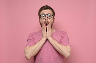 Gözlüklü ve pembe tişörtlü güzel bir adam şaşırır, elleriyle yüzünü tutar ve ağzı açık kameraya bakar..