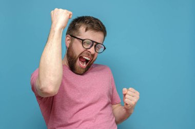 Gözlüklü ve pembe tişörtlü duygusal sakallı bir adam kazanan bir jest yapar ve neşeyle bağırır. Boşluğu kopyala.