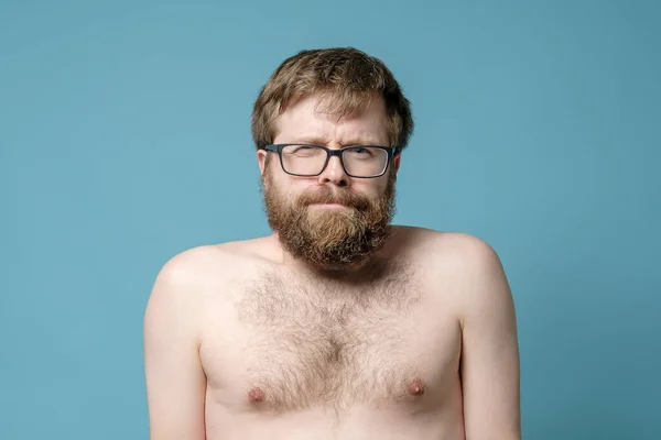Uomo divertente in occhiali con una barba arruffata e un'acconciatura troppo cresciuta, con un busto nudo, fissa la fotocamera, strizzando un occhio . — Foto Stock
