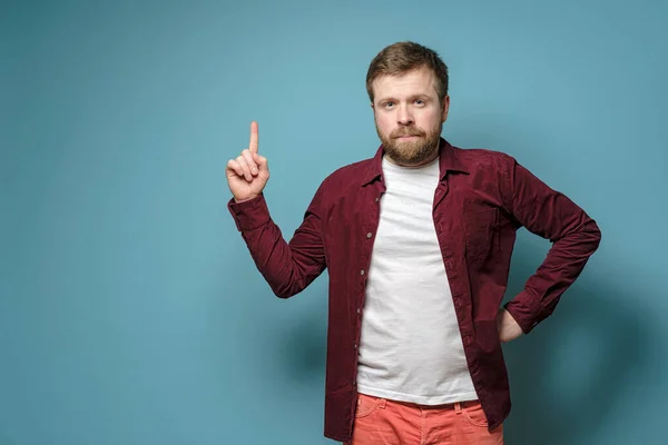 Calm, confident man shows index finger up and looks at the camera, on a blue background. Copy space. — Stock Photo, Image