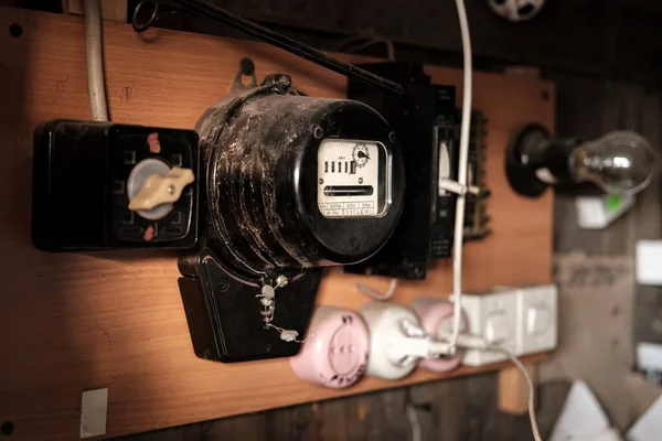 Old electric meter for calculating energy consumption is hanging in the barn, next to sockets and a light bulb. Russia. — Stock Photo, Image