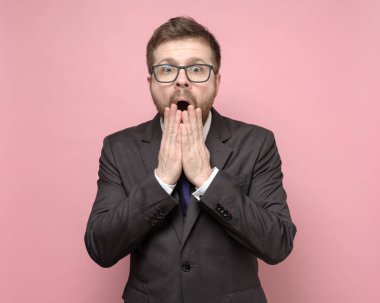 Gözlüklü ve takım elbiseli bir adam kameraya büyük gözlerle bakar ve ağzını elleriyle kapatır..