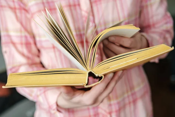 Nyílt vastag, régi könyv sárga színű női kéz, a háttérben egy rózsaszín kockás ing. — Stock Fotó