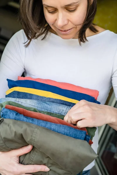 Mulher com uma mão segura calças e jeans cuidadosamente dobrados em cores brilhantes, e com o outro braço escolhe o que vestir . — Fotografia de Stock