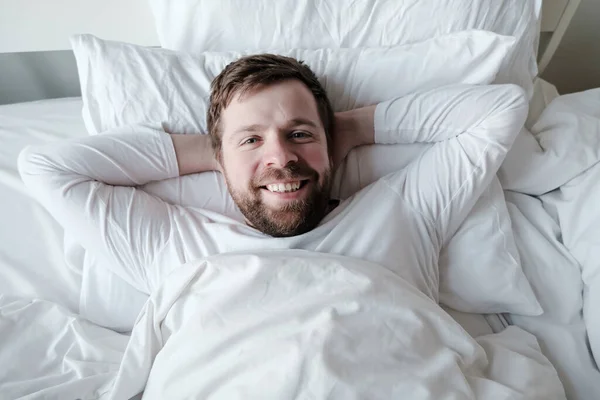 Nöjd man i pyjamas ligger i sängen på en mjuk kudde med händerna under huvudet, han är glad och ler. — Stockfoto