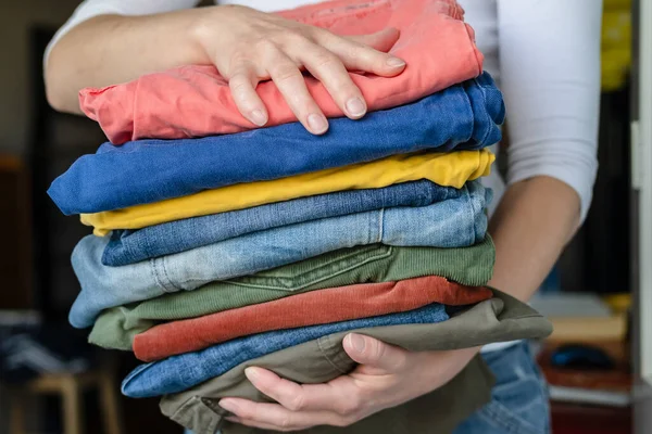 As mãos femininas mantêm calças e calças jeans bem dobradas em cores brilhantes, após a lavagem, em um fundo embaçado. . — Fotografia de Stock