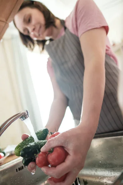 Female hands wash cucumbers and red tomatoes under stream water in a sink. Cute woman in an apron is preparing for cooking.