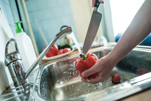 Weibliche Hand hält roten Pfeffer unter einem Wasserstrahl und wird ihn mit einem Messer schneiden, in der Küche. Essen kochen. — Stockfoto