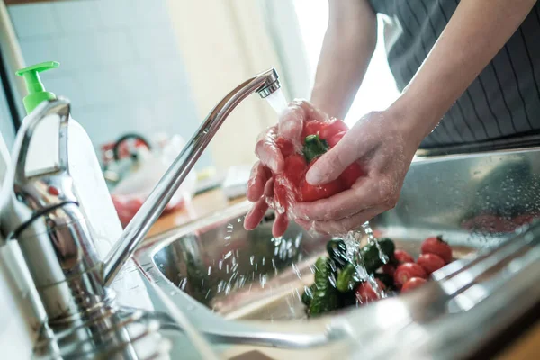 As mãos femininas lavam o pimentão vermelho abaixo de uma corrente de água, contra o contexto de verduras na pia. Cozinhar alimentos . — Fotografia de Stock