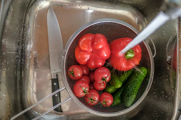 Fresh vegetables under a stream of water in a colander, next to a knife in the sink. Top view. — Stock Photo, Image