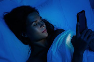 Genç ve güzel bir kadın gece yatakta akıllı telefon kullanıyor.