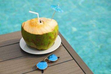 Yüzme havuzunun yanındaki masada taze hindistan cevizi içeceği ve güneş gözlüğü var.