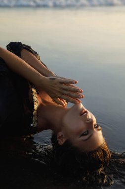 Siyah tunik giyen seksi ve ıslak kadın kumsalda siyah kumla poz veriyor.