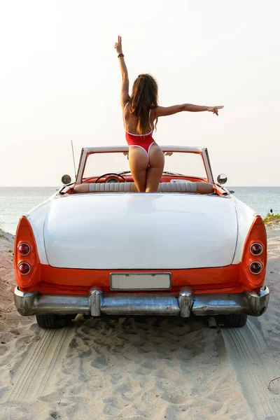 赤い水着を着たセクシー女性はビーチでレトロなキャブリオレット車の中に座っています — ストック写真