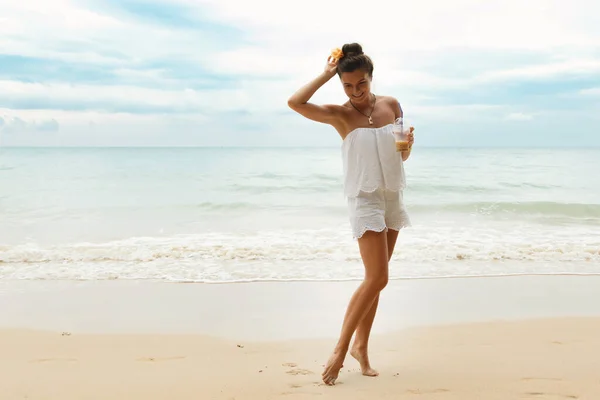 白いジャンプスーツを着た美しい女性氷のコーヒーを飲みながらビーチを歩く — ストック写真
