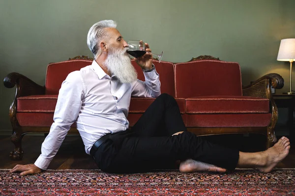 Stylish and handsome bearded senior man sitting on the floor and drinking red wine