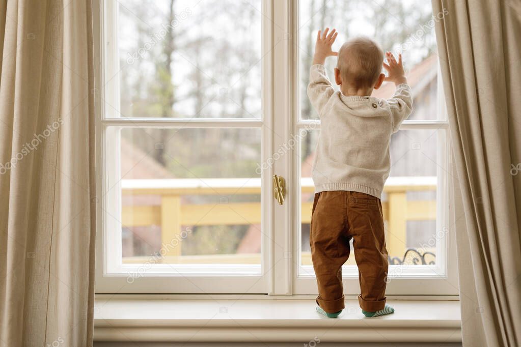 Little baby boy is standing on the windowsill and looking at the window