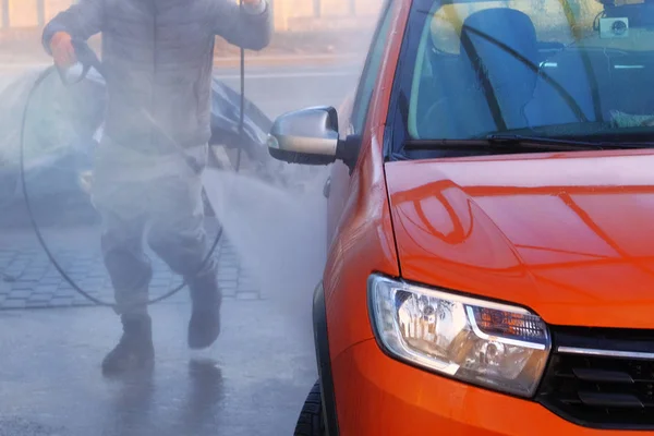 Man washes his orange car at car wash in outdoors. Cleaning with with a water jet at self-service car wash. Soapy water runs down.