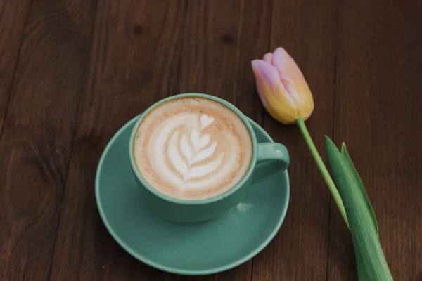 Coffee in a green cup with a pink tulip on a wooden background. Flat lay