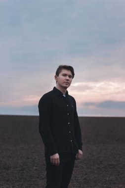 Sahada ciddi bir adam, siyah giyinmiş. Pembe gün batımı