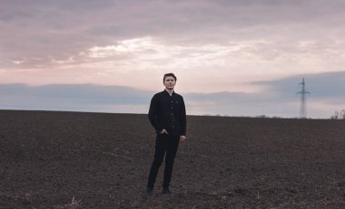 Sahada ciddi bir adam, siyah giyinmiş. Pembe gün batımı