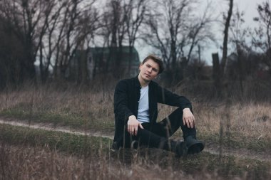 Portrait of a confident man sitting on a field. Thoughtful and successful. Black clothes. Field with dry grass. Vacation. Emotional exhaustion