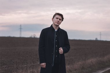 Sahada ciddi bir adam, siyah giyinmiş. Pembe gün batımı