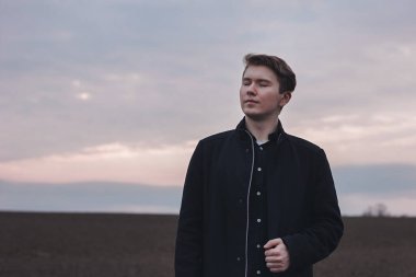 Sahada ciddi bir adam, siyah giyinmiş. Pembe gün batımı