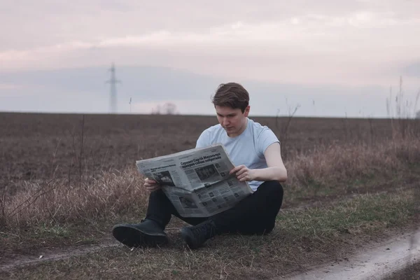 A man in a white T-shirt reads a newspaper in a field. Outdoor recreation