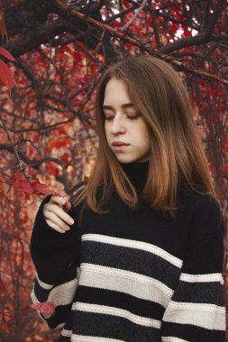 Genç bir kadın, serin bir sonbahar gününde çiçeklere nazikçe dokunurken kırmızı yaprakların arasında duruyor.