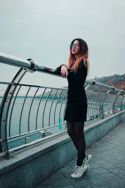 Pensive business woman looks into the distance on the background of the river and the bridge. Girl dressed in black and glasses. Business style. Woman with red lipstick