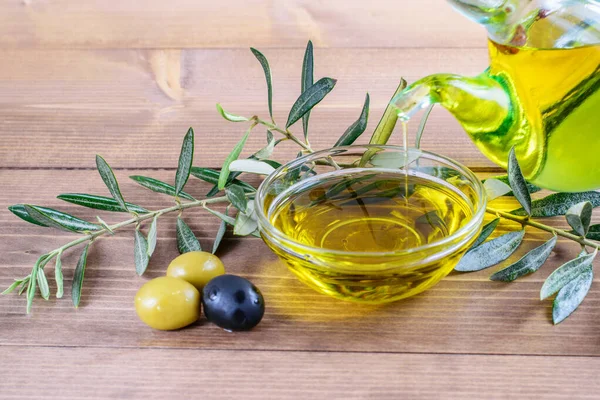 Cam şeffaf kâseye zeytin yağı damlıyor. Ahşap zemin üzerinde biraz zeytin ve zeytin dalı dalları. Sağlıklı gıda konsepti. 