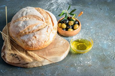 Bir somun yuvarlak beyaz ekmek, tahta kesme tahtasının üzerinde buğday kulakları. Koyu arkaplanda zeytin yağı ve ahşap kasede zeytin yağı. Ev yapımı fırın.. 