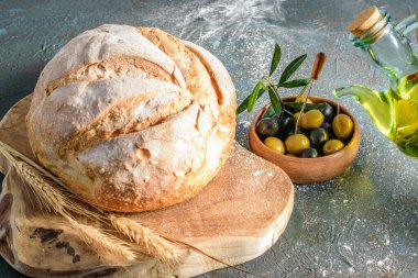 Beyaz yuvarlak ev yapımı ekmek ve mutfak tahtasının üzerinde biraz buğday kulağı. Ahşap kasede zeytin yağı şişesinde zeytin. Doğal sağlıklı yiyecekler. 
