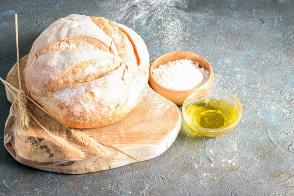 Handmade white bread and wheat ears on the wooden kitchen cutting board. White flour in wood bowl and olive oil on the dark background. Cooking natural handmade bread.