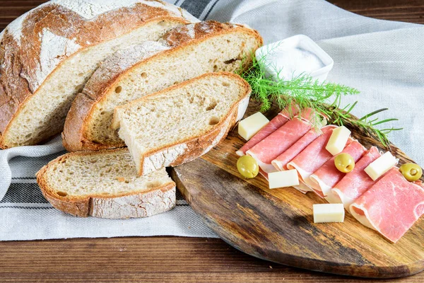 Tahta kesme tahtasında dilimlenmiş taze jamon, peynir ve zeytin. Mutfak peçetesinde ev yapımı integral ekmek ve tuz. Doğal ev yapımı sağlıklı gıda konsepti. 