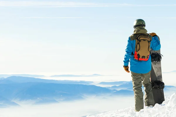 Joven snowboarder en pie casco al lado de snowboard cuenta en nieve con hermosos paisajes sobre fondo —  Fotos de Stock
