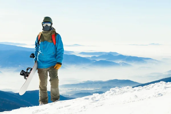 Close-up shot van jonge snowboarder met helm tegoed op de top van een berg met zijn snowboard in de hand — Stockfoto