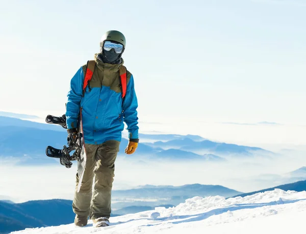 Jonge snowboarder in helm in de hand wandelen op de top van een berg met zijn snowboard — Stockfoto