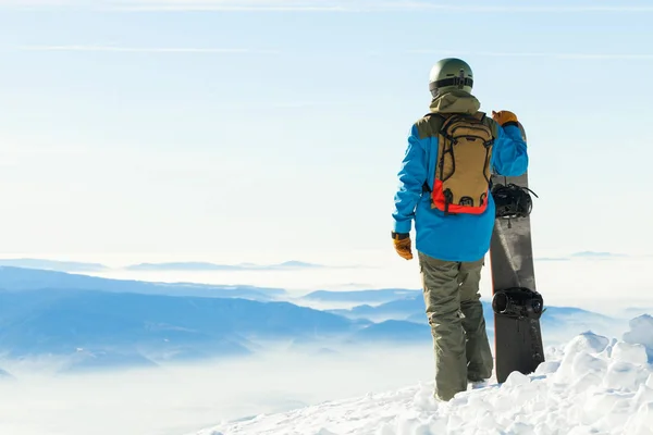 Snowboarder in helm permanent op de top van een berg en zijn snowboard met één hand te houden — Stockfoto