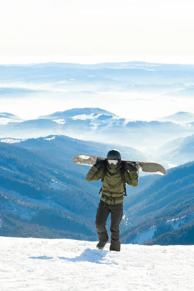 Snowboardzista młodych trzymając snowboard na jego sholders i chodzenie na sam szczyt góry śnieżne — Zdjęcie stockowe