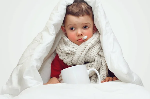 A little cute boy gets sick and measures the temperature with an oral thermometer and sits covered with a blanket and with a large knitted scarf around his neck.