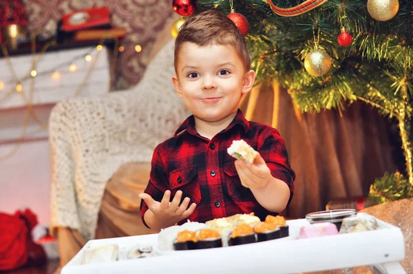 Cute boy eats sushi sitting under a Christmas tree in a red checkered shirt.